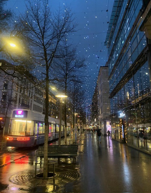 Long strands of twinkly white Christmas lights dangle above a wet street with buildings on either side and a tram running up the middle in early morning in Zurich, Switzerland.