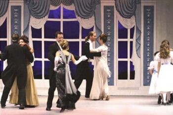 Stage scene for "The Sound of Music" with actors in formal attire dancing a choreographed waltz in front of a ballroom backdrop that feature a big picture window with white trim and blue billowy curtains.