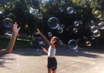 A teenage girl reaches for bubbles in a park while simultaneously holding her cell phone. It's modern multi-tasking!